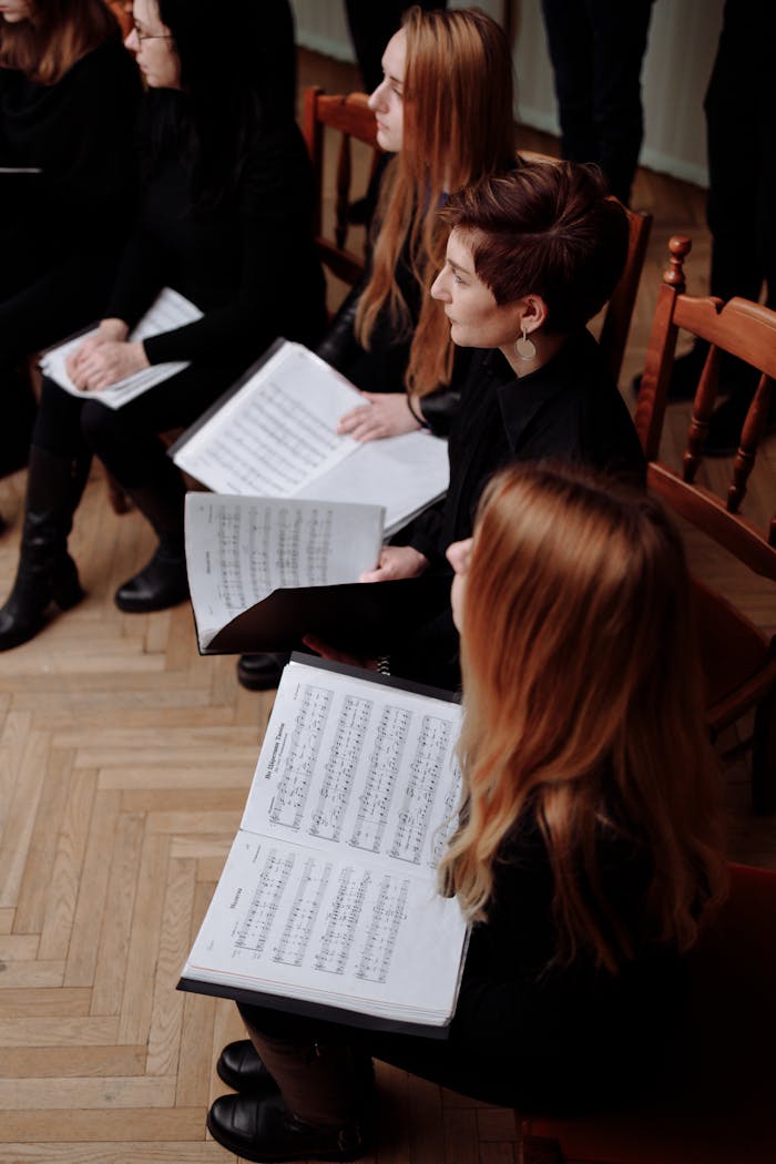 services-01 Group of women in a choir holding music sheets, seated on wooden chairs, practicing indoors.