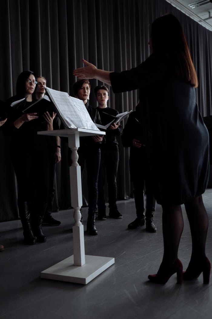 hero-img-02 A choir rehearses with a conductor standing by a music stand indoors, all in black attire.