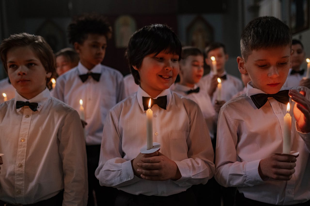 gallery-1 Children in formal white shirts holding candles during an indoor choir performance, fostering togetherness.